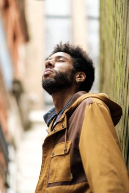 Sakallı ve kahverengi ceketli Afrikalı genç bir adam, bir duvara yaslanmış, dar bir sokakta gözleri kapalı derin bir nefes alıyor, temiz havanın ve farkındalığın tadını çıkarıyor.