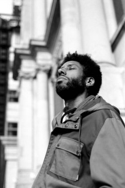 Kapalı gözlü, derin derin nefes alan ve şehir ortamında temiz havanın tadını çıkaran, huzur ve sükunet duygusu taşıyan genç bir adamın taneli monokrom analog fotoğrafı.