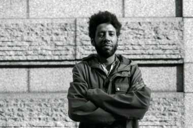 Taştan bir duvarın önünde kollarını kavuşturmuş, sıradan bir ceket giymiş ve ciddi görünen Afro-Amerikan genç bir adamın tek renkli analog fotoğrafı.