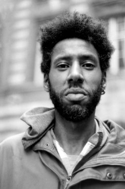 Afro saçlı ve sakallı, yüzünde ciddi bir ifadeyle poz veren Afro Amerikalı genç bir adamı gösteren 35 mm 'lik film kamerasından tek renk analog fotoğraf.