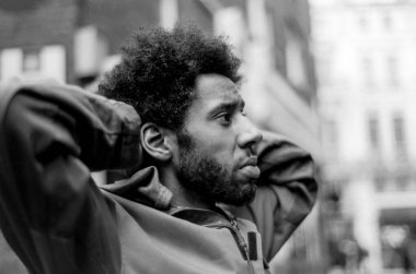 Mimari ve şehir hayatı ile dolu bulanık kentsel bir çevre ile çevrili, elleri başının arkasında gevşemiş afro saçlı genç bir adamı yakalayan taneli monokrom analog fotoğraf.