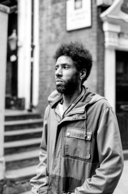 Kapüşonlu ceket giyen afro saçlı ve sakallı genç adam şehir ortamında duruyor. Kumlu, siyah ve beyaz bir anestetik fotoğrafta geçiyor.