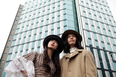 Şık şapkalar giyen şık erkek ve kadın modern bir gökdelene bakıyor, kentsel mimariyi canlı bir şehir manzarasıyla modern modayla harmanlıyor.