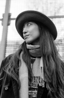 Taneli tek renkli analog fotoğraf şık bir kadife ceket, şapka ve eşarp giymiş genç bir kadını kentsel bir arkaplan ortasında düşünceli bir ifadeyle yukarı bakarken yakalıyor.