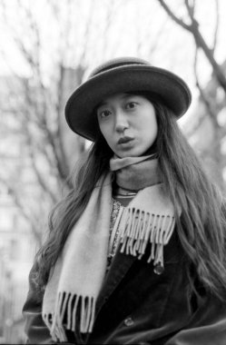 Tahıllı, tek renkli, analog tarzda, şapka ve eşarp takan genç bir Asyalı kadının portresi, çıplak ağaçların bulanık arka planına poz vermek, klasik, filmden esinlenilmiş bir estetiği çağrıştırıyor