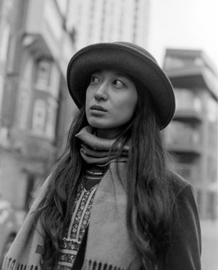 Grainy monokrom analog vinog fotoğraf, şapka ve eşarp takan şık bir genç kadını çekiyor. Kalabalık şehir sokaklarında yürürken yukarı bakıyor. Zarafet ve merakın vücut bulmuş hali.