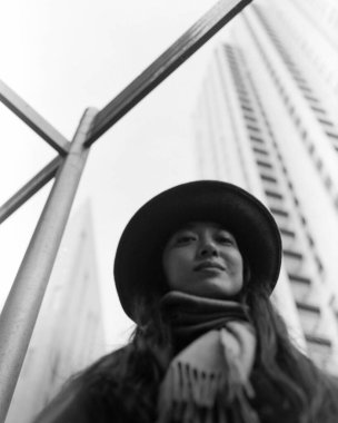 Siyah ve beyaz analog fotoğraf, şapka ve eşarp takmış şık bir kadın, çarpıcı metal yapılar ve gökdelenlerin kentsel arkaplanlarına karşı güvenle poz veriyor