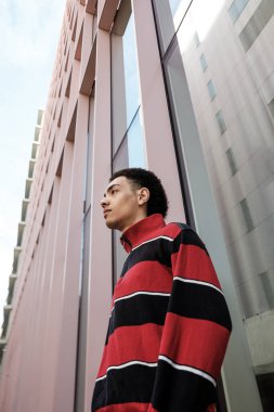 Şık bir genç adamın çizgili bir kazak giymesi, canlı bir şehir ortamında modern bir binanın yanında dikilip izlemesi, modaya uygun bir yaşam tarzını yakalaması düşük açılı bir bakış açısı.