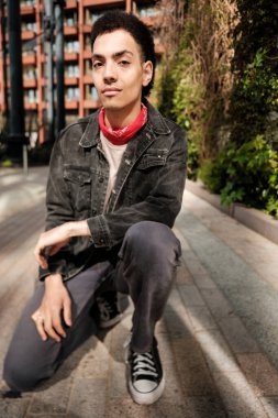 Bandanalı şık bir genç adam şehirde diz çöküp poz veriyor, modern şehir moda eğilimlerini somutlaştırıyor. Bir şehir zeminine karşı çağdaş gençlik kültürünün özünü yakalamak