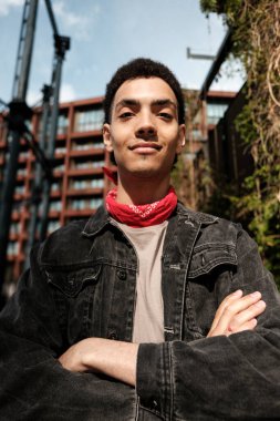 Kırmızı bandana takan şık bir genç adam ve kot ceketi kollarını kavuşturarak modern bir şehir zeminine karşı güven ve serinlik yayıyor.