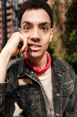 Kırmızı bandanalı ve siyah kot ceketli genç adam çenesine dokunurken komik bir yüz ifadesi takınıyor.