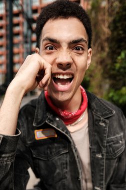 Bandana ve kot ceket giyen şık genç adam, dışarıda poz verirken komik bir yüzle bağırıyor, canlı bir şehir ortamında heyecan ve neşe saçıyor.