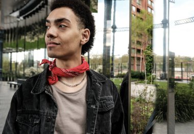 Kırmızı bandana ve kot ceket giyen şık genç adam, modern bir şehir ortamında kendine güvenerek duruyor, güncel moda eğilimlerini ve genç sokak tarzını yansıtıyor.