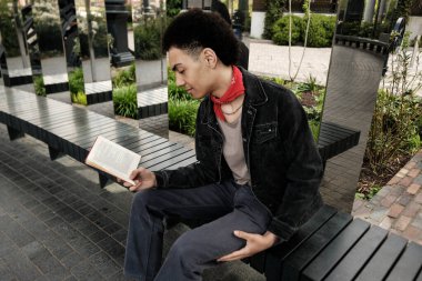 Genç bir öğrenci, çağdaş bir halk parkında, yansıtıcı sütunlarla süslenmiş bir bankta otururken kitap okumaktan zevk alıyor, edebiyata dalmış, huzurlu bir şehir ortamının ortasında.