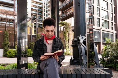 Genç adam modern bir şehir parkında bir bankta oturuyor, bir kitap okumaya dalmış, kentsel çevrede huzurlu bir anın tadını çıkarıyor.