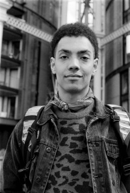 Taneli monokrom analog film portresi kot ceket giyen genç bir adam, desenli kazak ve bandana, modern bir şehri bir sırt çantasıyla keşfediyor, klasik bir estetik sergiliyor.