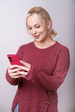 Kestane rengi kazak giyen neşeli sarışın kadın kırmızı bir cep telefonu kullanıyor, internette geziniyor ya da online iletişimden hoşlanıyor, stüdyo arka planında izole edilmiş.