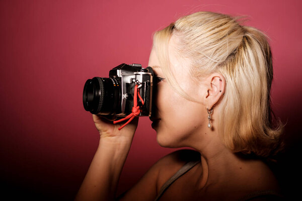 Young blonde photographer taking pictures using a vintage analogic camera with a red strap, concentrating on framing the perfect shot on a vibrant pink background