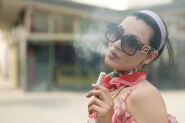 Şehir ortamında güneş gözlüğü ve modaya uygun kıyafetler giyerken vapurdan duman üfleyerek rahatlama anının tadını çıkaran modaya uygun bir etkili.