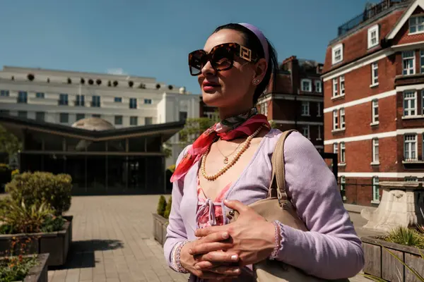 Güneş gözlüğü, inci kolye ve şık kıyafetler takan şık bir kadın açık havada poz veriyor. Şık bir şehir ortamında zarafet ve şehir şıklığını sergiliyor.