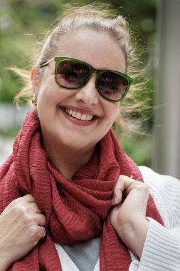 Neşeli, olgun bir kadın güneş gözlüğü takıyor ve canlı kırmızı bir eşarp açık havada sıcacık gülümsüyor, kendine güven yayıyor ve şık, rahat bir ortamda hayat dolu bir ruh.