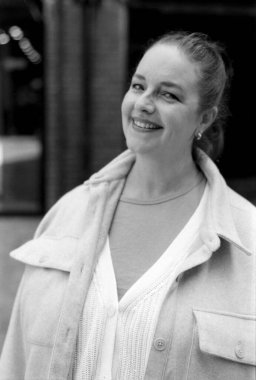 35 mm 'lik film kamerasından alınan tek renkli kareli analog fotoğrafta açık renk ceket giyen ve arka planı bulanık bir hırka giyen gülümseyen bir kadın görülüyor.