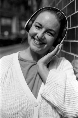 Kablosuz kulaklık takan, gülümseyen bir kadının, onlara eliyle dokunduğu ve tuğla bir duvara yaslandığı, kentsel bir ortamda müziğin keyfini çıkardığı tek renkli, tek renkli, klasik bir fotoğraf.