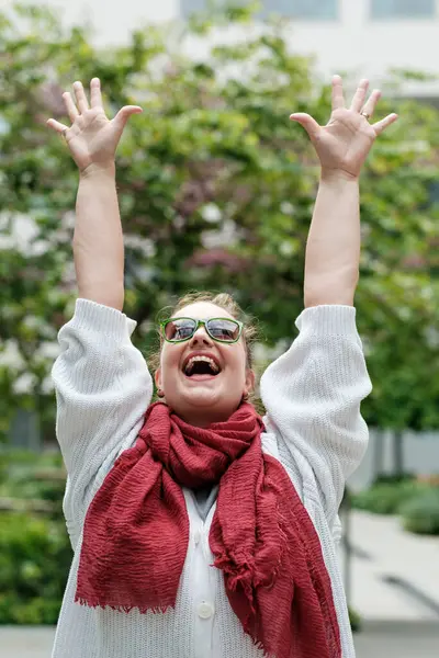 Heyecanlı, güneş gözlüğü takan, kırmızı eşarp takan mutlu kollarını kaldıran ve dışarıda başarıyı kutlayan mutlu ve özgür bir kadın.