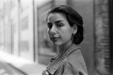 Üzerinde kot ceket ve küçük küpeler olan ve arka planda bulanık binalar bulunan şehir ortamında duran 35 mm 'lik tek renkli bir fotoğrafçı portresi.