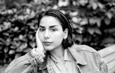 Tahıllı, siyah ve beyaz, analog fotoğraf kısa saçlı, kot ceketli ve polo gömlekli genç bir kadını çekiyor, yüzünü eline yaslıyor, klasik, düşünceli bir hava yaratıyor.