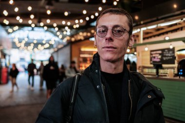 Gözlüklü şık bir adam ve sırt çantalı siyah bir ceket modern bir gıda pazarında poz veriyor. Arka planda yürüyen insanlar canlı ve kentsel bir atmosfer yaratıyor.