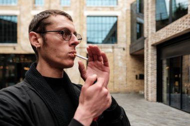 Gözlüklü ve küpeli genç bir adam sigarasını çakmakla yakıyor. Tuğladan binalarla dolu modern bir şehir ortamının arka planında duruyor.