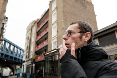 Genç adam sigara içerken Londra caddesinde yürürken, demiryolu köprüsü ve klasik tuğla binalar bulutlu bir günde bir zemin oluşturur.