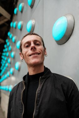 Siyah balıkçı yaka kazak ve bombardıman ceketi giyerken gülümseyen şık bir adam, mavi parlak ışıklarla süslenmiş fütürist bir duvarın yanında güvenle duruyor.