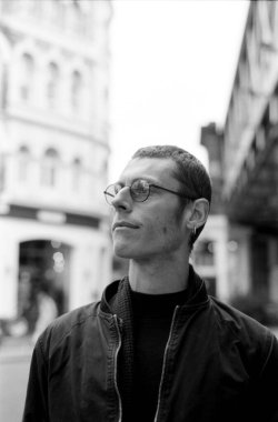 Yuvarlak gözlüklü ve siyah ceketli bir gencin, arka planda bulanık bir bina ve yükseltilmiş bir demiryolu olan bir şehir caddesinde dikilirken çekilmiş tek renkli analog fotoğrafı.