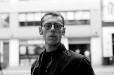 Gözlük ve küpe takan, şehir ortamında poz veren, biraz bulanık arka planlı, görüntüye klasik bir film estetiği veren tek renkli bir genç adam.