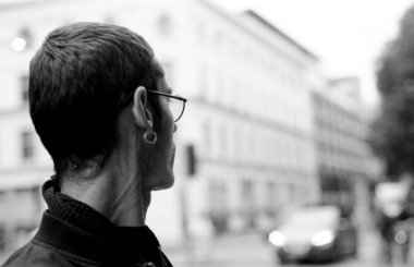 Gözlüklü ve küpeli bir adamın arka planda bulanık binaları ve arabası olan bir şehir caddesini düşünürken çekilmiş tek renkli analog fotoğrafı. 35 mm 'lik film estetiğini anımsatıyor.