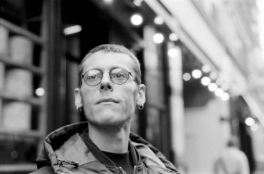 Siyah beyaz analog film portresi. Yuvarlak gözlüklü ve kapüşonlu ceketli genç bir adam. Bulanık bir şehir arka planına kurulmuş.