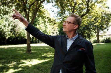 Gülümseyen kıdemli profesör, parkta dikilirken sağ işaret parmağıyla gökyüzündeki bir şeyi işaret ederek öğrencilerine muhtemelen bir kavramı anlatıyor.