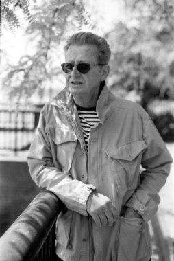 Grainy monokrom analog film fotoğrafı güneş gözlüğü takan ve açık renk ceket giyen bir adamın bir eli cebinde korkuluklara yaslanarak klasik bir estetiği anımsattığını gösteriyor.