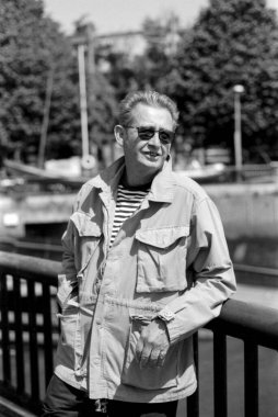 Yaşlı bir adamın güneş gözlüğü taktığı ve parktaki korkuluklara dayanan açık renk ceketli, bulanık ağaçlı ve arka planda bir köprü olan tek renkli, klasik bir fotoğraf.