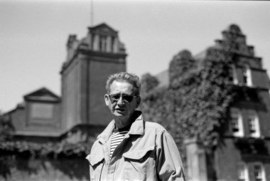 Yaşlı bir adamın güneş gözlüğü taktığı ve eski, kısmen sarmaşık kaplı bir binanın önünde bir kuleyle poz verdiği tek renkli, klasik bir portre.