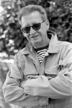 Güneş gözlüğü takan ve kolları bağlı açık ceketli gülümseyen tek renkli, analog bir film portresi. Açık havada güneşli bir gün ve bulanık bir arka plan.