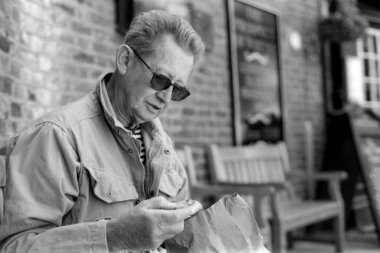 Taneli monokrom görüntüsü, güneş gözlüğü ve günlük giysiler takan son sınıf bir adamı dışarıda bir barda otururken kese kağıdından bir şeyler yerken yakalıyor.