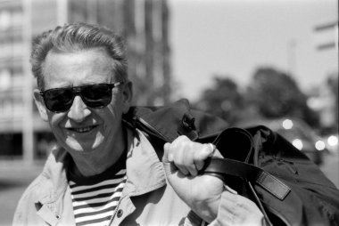 Güneş gözlüğü takan ve güneşli bir şehir ortamında omzunda siyah bir çanta taşıyan gülümseyen yaşlı bir adamı gösteren tek renkli, klasik bir fotoğraf.