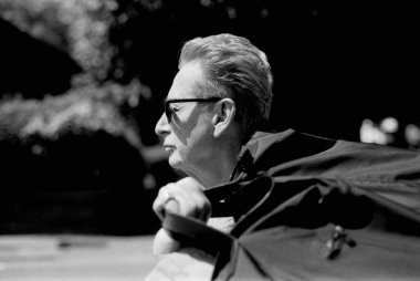 Yaşlı bir adamın güneş gözlüğü taktığı ve ceketini tutup profil görüntüsünde kameradan yüzünü çevirdiği tek renkli, klasik bir fotoğraf.