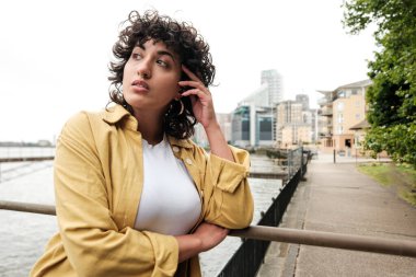 Kıvırcık saçlı ve sarı ceketli genç bir kadın şehirdeki bir nehrin kenarında duruyor, korkuluklara yaslanıyor ve düşünceli bir şekilde uzaklara bakıyor, geleceğini düşünüyor.