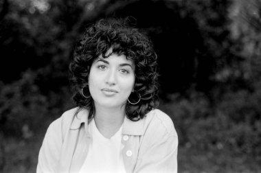 Kıvırcık saçlı, halka küpeli, beyaz bir tişörtün üzerine açık renk bir ceket giyen ve hafifçe bulanık, doğal bir fonda poz veren genç bir kadının tek renkli portresi.