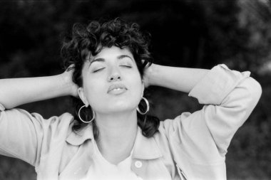 Grady monokrom analog eski bir fotoğraf, kıvırcık saçlı, küpe takan ve açık renk ceketli, ellerini başının arkasında tutan, gözleri kapalı ve gevşek bir ifadeyle genç bir kadını gösteriyor.