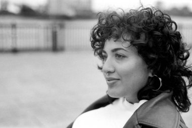 Tahıllı tek renkli portre kıvırcık saçlı, burnu delinmiş, şık giyinmiş, arka planda bulanık poz veren genç bir kadını çekiyor.